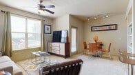 Located at 18211 Bulverde Rd in San Antonio, Texas, this single-floor residence offers a thoughtfully designed layout that balances comfort and functionality. The open-concept living and dining area is filled with natural light from large windows dressed with draping curtains and adjustable blinds. A ceiling fan overhead and plush carpeting underfoot add to the inviting atmosphere, while track lighting illuminates the adjacent dining space. The kitchen features rich dark cabinetry contrasted by granite-style countertops, black appliances, and wood-look flooring, providing a sleek and practical cooking environment. A dedicated laundry area with a full-size washer and dryer is conveniently accessible directly from the kitchen. The bedroom is carpeted and bathed in natural light through a window with sheer curtains, offering a peaceful retreat. A walk-in closet provides ample storage. The bathroom includes a full tub-shower combination with classic white tile accented by a dark tile border, complemented by dark cabinetry and granite countertops. Throughout the residence, neutral tones and consistent finishes create a cohesive and comfortable living environment well-suited for everyday life in the heart of San Antonio.