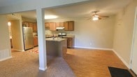 This single-floor apartment at Brookwood Apartments offers an open-concept design that seamlessly connects the kitchen and living area. The kitchen is well-appointed with warm-toned wooden cabinetry, granite-style countertops, and stainless steel appliances, including a refrigerator and electric range with an overhead ventilation hood. A peninsula with additional counter space serves as a natural divider between the kitchen and the adjoining living area, providing both extra prep space and an informal gathering spot. The living area features a combination of hardwood flooring and carpeting, clearly delineating the transition between spaces. A ceiling fan with an integrated light fixture provides both airflow and ambient lighting to the living area. Neutral gray walls throughout create a clean, cohesive aesthetic, while white trim and structural columns add subtle architectural definition. The layout maximizes the sense of openness, making the space feel inviting and functional. Overall, the apartment delivers a practical, comfortable living environment with a straightforward, modern design.