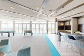 Located at 1360 West University Avenue in Gainesville, FL, this single-floor amenity lounge at The Standard offers a generously sized, open-concept recreational space designed for socializing and entertainment. The interior features a contemporary design with a coffered ceiling, recessed lighting, and decorative pendant fixtures that add warmth to the modern aesthetic. A vivid turquoise carpet runner bisects the light tile and neutral carpet flooring, providing a striking visual accent throughout the space. The lounge is equipped with a full-length bar counter finished with mosaic tile accents and lined with sleek bar stools, ideal for casual gatherings. A built-in seating nook with cabinetry and a dining area offers additional space for relaxed interaction. Recreation amenities include a regulation billiards table and a foosball table, catering to a variety of leisure activities. Floor-to-ceiling glass walls and oversized sliding glass doors flood the interior with abundant natural light and open directly onto an outdoor terrace, providing expansive panoramic views of the surrounding Gainesville skyline. A bold green living wall branded with "The Standard" logo serves as a distinctive focal point near the entrance, reinforcing the space's upscale character and identity.