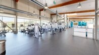 Located at 1580 Jade St in Davis, CA, this single-floor fitness facility offers a generously sized exercise room designed to accommodate a wide range of workout needs. The space is characterized by soaring ceilings with exposed warm-toned wood beam accents and industrial-style ductwork, lending the interior a modern, open aesthetic. Large floor-to-ceiling windows line the walls throughout, flooding the facility with abundant natural light and offering views of the surrounding outdoor areas, including glimpses of a pool.

The main exercise floor is finished with durable rubber flooring and is organized into distinct workout zones. A comprehensive cardio section features rows of treadmills, ellipticals, and upright bikes positioned to take advantage of the natural light and outdoor views. The strength training area is well-equipped with a full array of selectorized machines, free-weight benches, and multiple dumbbell racks backed by mirrored walls. A dedicated functional training zone provides open floor space alongside TRX suspension trainers, medicine balls, and balance equipment. A vibrant teal accent wall adds a bold pop of color, enhancing the energetic atmosphere. The facility's thoughtful layout and high-end equipment make it a comprehensive destination for fitness.
