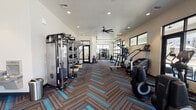 Located in North Phoenix at North Valley Pkwy, this single-floor fitness center is designed to support a comprehensive workout experience. The space features a striking herringbone-patterned carpet in brown, gray, and teal tones that runs throughout, lending a modern and cohesive aesthetic to the entire facility.

The cardio zone is generously equipped with a row of treadmills, elliptical trainers, and a stair climber, all positioned along floor-to-ceiling windows that offer views of the surrounding outdoor amenity areas, filling the space with abundant natural light. Large-blade ceiling fans are mounted throughout, promoting airflow and comfort during workouts.

The strength training area includes a multi-functional cable and free-weight machine, adjustable benches, and additional resistance equipment. Mirrored walls in key zones enhance the sense of space and allow users to monitor their form. A glass-panel garage door opens directly to an outdoor turf area, seamlessly connecting the interior training space to the exterior and allowing for fresh-air workouts.

Additional amenities include a water fountain, a storage cubby unit for personal belongings, and a wall-mounted television. Recessed lighting and multiple windows ensure the space remains bright and well-lit throughout the day. The overall design is clean, functional, and well-appointed for residents seeking a high-quality fitness facility.
