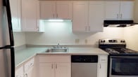 This single-floor space includes a well-appointed kitchen designed with a clean, cohesive aesthetic. The kitchen features an abundance of white shaker-style cabinetry, offering generous storage both above and below the countertops. Light-toned counters run along the workspace, providing ample room for meal preparation. A stainless steel sink is centrally positioned beneath under-cabinet lighting that illuminates the work surface efficiently, contributing to the kitchen's bright and functional atmosphere. Appliances include a black electric range and a stainless steel dishwasher, delivering practical, everyday convenience. The white walls and cabinetry work together to create a clean, open feel throughout the kitchen. The consistent use of white tones and silver hardware lends the space a timeless, neutral character that is easy to personalize. Overall, the kitchen is organized and practical, well-suited for everyday cooking and entertaining needs within this single-story layout.