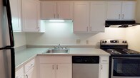This single-floor space includes a well-appointed kitchen designed with a clean, cohesive aesthetic. The kitchen features an abundance of white shaker-style cabinetry, offering generous storage both above and below the countertops. Light-toned counters run along the workspace, providing ample room for meal preparation. A stainless steel sink is centrally positioned beneath under-cabinet lighting that illuminates the work surface efficiently, contributing to the kitchen's bright and functional atmosphere. Appliances include a black electric range and a stainless steel dishwasher, delivering practical, everyday convenience. The white walls and cabinetry work together to create a clean, open feel throughout the kitchen. The consistent use of white tones and silver hardware lends the space a timeless, neutral character that is easy to personalize. Overall, the kitchen is organized and practical, well-suited for everyday cooking and entertaining needs within this single-story layout.