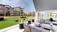 This expansive outdoor amenity space is designed to foster community and leisure within a modern residential complex. At its centerpiece stands a striking covered pavilion with a soaring vaulted ceiling clad in corrugated metal, supported by stone-accented columns. The pavilion houses a dramatic double-sided stone fireplace that anchors the space, providing warmth and visual appeal. Floor-to-ceiling glass walls and dark-framed double doors allow seamless transitions between the covered interior and the open-air courtyard.

The pavilion's sheltered patio is furnished with contemporary seating arrangements — including plush sofas, accent chairs, and cylindrical side tables — alongside mounted flat-screen televisions for outdoor entertainment. String lights drape across the courtyard, creating a warm and inviting ambiance day and evening.

Beyond the pavilion, a generously sized artificial turf lawn serves as the hub for outdoor recreation, accommodating cornhole boards and casual gatherings. Wicker lounge chairs and umbrella-shaded seating clusters are scattered throughout, offering additional gathering spots. The surrounding multi-story residential buildings, constructed with a blend of brick, stone, and contemporary paneling, frame the courtyard with a cohesive architectural character. Manicured native landscaping and pathway lighting complete the polished, resort-style outdoor experience.