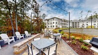 Located at 11023 Moonbug Ct in Charlotte, North Carolina, this outdoor amenity space is thoughtfully designed to offer residents a well-appointed al fresco gathering environment. The centerpiece of the area is a generously sized paver patio finished in warm-toned brick, accommodating multiple dining tables and chairs ideal for casual meals or social gatherings. Overhead, a canopy of string lights is strung between tall poles, creating an inviting atmosphere during evening hours.

Several gas grilling stations are positioned throughout the patio, providing convenient outdoor cooking options. Along the perimeter, white Adirondack chairs are arranged on a separate concrete seating pad, offering a relaxed vantage point overlooking the landscaped grounds. Neatly mulched planting beds with young shrubs and greenery border the space, adding natural texture and color.

A decorative iron fence lines the edge of the property, beyond which a scenic backdrop of mature deciduous trees—displaying vibrant autumn foliage—frames a tranquil wooded view. The surrounding multi-story residential buildings are finished in crisp white and gray exteriors, complementing the clean, modern aesthetic of the outdoor space. The overall ambiance is one of a well-maintained, community-focused retreat that seamlessly blends structured amenities with the natural beauty of the Charlotte landscape.