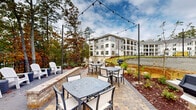 Located at 11023 Moonbug Ct in Charlotte, North Carolina, this outdoor amenity space is thoughtfully designed to offer residents a well-appointed al fresco gathering environment. The centerpiece of the area is a generously sized paver patio finished in warm-toned brick, accommodating multiple dining tables and chairs ideal for casual meals or social gatherings. Overhead, a canopy of string lights is strung between tall poles, creating an inviting atmosphere during evening hours.

Several gas grilling stations are positioned throughout the patio, providing convenient outdoor cooking options. Along the perimeter, white Adirondack chairs are arranged on a separate concrete seating pad, offering a relaxed vantage point overlooking the landscaped grounds. Neatly mulched planting beds with young shrubs and greenery border the space, adding natural texture and color.

A decorative iron fence lines the edge of the property, beyond which a scenic backdrop of mature deciduous trees—displaying vibrant autumn foliage—frames a tranquil wooded view. The surrounding multi-story residential buildings are finished in crisp white and gray exteriors, complementing the clean, modern aesthetic of the outdoor space. The overall ambiance is one of a well-maintained, community-focused retreat that seamlessly blends structured amenities with the natural beauty of the Charlotte landscape.