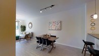 This thoughtfully designed single-floor space offers a seamless open-plan layout that combines a dining area and living space within a bright, welcoming environment. The dining area is anchored by a solid wood table paired with sleek black chairs with chrome frames, creating a stylish yet functional gathering spot. A large, colorful abstract artwork adorns the wall above, adding a vibrant focal point to the otherwise neutral palette. Track lighting installed on the ceiling provides adjustable, directional illumination throughout the space. Light-toned hardwood flooring runs throughout, lending warmth and continuity to the interior. The adjacent living area is partially visible and features a comfortable sofa, a woven accent chair, and layered textiles that contribute to a relaxed ambiance. A decorative sunburst mirror and lush indoor greenery add organic, textural interest to the walls and corners. The clean white walls and high, smooth ceilings amplify the sense of openness and allow natural light to reflect freely across the space. The overall atmosphere is modern and livable, with a balanced mix of contemporary furnishings and decorative accents that create a cohesive, inviting interior.