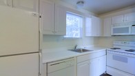 This single-floor space offers a functional and well-maintained layout designed for comfortable everyday living. The kitchen is a standout feature, finished in a clean, all-white palette that creates a bright and airy atmosphere. White upper and lower cabinetry provides generous storage, complemented by expansive white countertops that offer ample workspace for meal preparation. A stainless steel sink is conveniently positioned beneath a window fitted with blinds, allowing natural light to fill the room while offering a view of the surrounding greenery outdoors. White appliances, including a full-size refrigerator and a dishwasher, are seamlessly integrated into the kitchen's cohesive design. Flush-mount ceiling lighting ensures the space remains well-lit throughout the day. The overall aesthetic is simple, clean, and practical, making efficient use of the available space. The single-floor layout contributes to an accessible and straightforward flow throughout the home, making it well-suited for a variety of occupants. The consistent white finish across cabinetry and appliances lends the kitchen a timeless, neutral character that is easy to personalize.