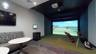 This single-level indoor golf simulator space is thoughtfully designed for both serious practice and recreational play. The room is defined by a large, floor-to-ceiling projection screen that dominates one wall, displaying vivid, lifelike golf course imagery to create an immersive playing environment. Overhead, a ceiling-mounted projector delivers sharp, high-quality visuals, while recessed and track lighting illuminate the space effectively without interfering with gameplay.

The hitting area features artificial turf matting that simulates a realistic fairway surface, providing a comfortable and functional stance for golfers of all skill levels. A dedicated computing station with a wall-mounted monitor allows players to manage simulator settings, track performance data, and select from a variety of virtual courses.

The surrounding walls are finished in a clean grey tone, contrasting with the dark ceiling treatment that gives the simulator bay a focused, cave-like ambiance suited for screen visibility. A patterned area rug and a small accent table at the periphery provide a subtle lounge element, making the space functional for both active participants and observers. The overall design balances technical functionality with a polished, modern aesthetic.