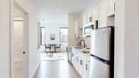 Located at 100 N Farwell St in Eau Claire, Wisconsin, this efficiently designed studio offers a thoughtful single-floor layout that maximizes every square foot. The open-concept kitchen and living area serves as the heart of the unit, featuring a galley-style kitchen outfitted with white shaker cabinetry with matte black hardware, a subway tile backsplash, stainless steel appliances including a refrigerator, microwave, and dishwasher, and a undermount sink with a black faucet. Light wood-tone flooring runs throughout, complementing the bright white walls and recessed lighting to create a clean, contemporary aesthetic. The living area accommodates a Murphy bed with built-in wood shelving above, offering flexible use of the space, alongside a seating area bathed in natural light from large, modern windows with views of the surrounding Eau Claire cityscape. A dedicated entryway nook includes a built-in bench and overhead cabinetry with wall hooks, providing practical storage upon entry. The full bathroom features tile flooring, a wood-toned vanity with a white countertop, a large mirror, and a shower-tub combination. A separate laundry closet houses a full-size front-loading washer. The overall ambiance is modern, clean, and efficiently planned.