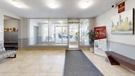 Located at 221 Glenridge Ave in St. Catharines, Ontario, this building lobby serves as the welcoming entrance to a Homestead residential property. The ground-floor space is finished with light beige ceramic tile flooring accented by grey carpet runners, creating a clean and functional aesthetic throughout.

Upon entry, floor-to-ceiling glass panels and automatic doors flood the lobby with natural light while offering clear sightlines to the exterior parking area. Flush-mounted ceiling light fixtures provide consistent ambient illumination across the space.

Moving inward, a pair of stainless steel elevators set against a stone-textured wall serve as the primary vertical access point for residents. A resident information board is mounted nearby, offering building notices and updates. Adjacent to the elevators, a glass-panel door provides access to the main residential corridor.

A comfortable seating area is positioned toward the interior, furnished with two upholstered armchairs and a small side table, alongside a framed abstract artwork that adds a touch of character to the neutral walls. A wooden bench near the elevator bank offers additional seating. Security is supported by a visible surveillance camera.

The lobby also connects to a secondary corridor, with a mailbox area visible through a side passage. Overall, the space presents a practical, well-maintained, and welcoming environment for residents and visitors alike.