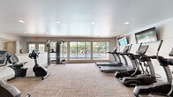 This fitness center offers a comprehensive workout environment within a single, open-plan space finished with neutral carpet tiles and recessed lighting throughout. The room is thoughtfully organized into distinct workout zones to accommodate a variety of exercise routines.

The cardio area is anchored by a row of TRUE-brand treadmills, each paired with a dedicated wall-mounted flat-screen TV, allowing users to stay entertained during their workouts. An upright stationary bike and a recumbent bike provide additional low-impact cardio options. Large windows along one wall flood the space with natural light and offer views of an outdoor pool and patio area, creating an open and energizing atmosphere.

The strength and functional training zone features a multi-station cable machine, an adjustable workout bench, and a seated row machine, catering to a full range of resistance exercises. A dedicated free weights area includes a rack of dumbbells in varying weights, complemented by a selection of medicine balls and stability balls stored on a wall-mounted rack. The clean, white walls and neutral color palette throughout contribute to a bright and motivating environment. The space is accessible via a dedicated entry door and is equipped with emergency exit signage, reflecting a practical and safety-conscious design.