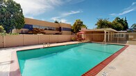 Located at 13475 Bancroft Avenue in San Leandro, California, this outdoor pool area serves as a shared amenity space within a residential community. The centerpiece is a generously sized rectangular swimming pool with vivid turquoise water, framed by a classic brick coping that adds a timeless architectural touch to the surroundings. The pool deck is finished in light-toned concrete, providing ample space for lounging and movement around the perimeter.

A dedicated seating area is positioned poolside, complete with tables and red umbrella shades that offer relief from the California sun. The space is fully enclosed by a combination of wooden privacy fencing and metal security fencing, with a clearly marked exit gate ensuring safe and controlled access. A life preserver ring is mounted on the fence, reflecting attention to safety standards.

Mature trees and well-kept shrubbery line the perimeter, contributing to a sense of privacy and natural comfort. The multi-story residential buildings that frame the background feature balconies and staircases that overlook the pool area, reinforcing the communal character of the space. Under open skies, the pool area benefits from abundant natural light, making it an inviting outdoor retreat for residents throughout the warmer months.