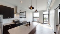 Located at 449 1/2 Bridge St NW in Grand Rapids, Michigan, this single-floor unit showcases a contemporary industrial design with thoughtful finishing details throughout. The open-concept kitchen serves as a centerpiece, featuring dark cabinetry, stainless steel appliances, a subway tile backsplash, a wall-mounted range hood, and distinctive black pipe shelving that adds an urban edge. A spacious kitchen island with a quartz countertop and undermount sink provides both prep space and casual dining opportunities, illuminated by stylish Edison bulb pendant lights overhead. Exposed ductwork adds to the industrial aesthetic. Light-toned hardwood-style flooring flows consistently throughout the unit, complementing the neutral gray walls and dark trim accents. A rustic barn door with black hardware provides a striking focal point and leads to the in-unit laundry area, blending function with design flair. The bedroom is bright and airy, benefiting from a large picture window that fills the room with natural light and offers views of the surrounding neighborhood. The bathroom is well-appointed with a tub-shower combination, matte black fixtures, a dark wood vanity, and a sliding glass door entry. Large windows in the main living area frame open city views, enhancing the sense of space and connecting the interior to the vibrant Grand Rapids surroundings.