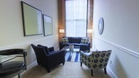 This two-floor space masterfully combines industrial character with refined, contemporary design. The living area immediately captures attention with its soaring ceilings that amplify the sense of openness and grandeur. Exposed brick accents frame large windows dressed with vertical blinds, allowing an abundance of natural light to pour in and illuminate the room's warm, neutral tones.

The seating area is thoughtfully arranged, featuring a navy blue sofa paired with coordinating armchairs — one in solid navy and another in a bold, patterned fabric — creating an inviting and stylishly curated atmosphere. A glass-topped coffee table anchors the space atop a striped area rug, while contemporary artwork adorns the walls, adding artistic flair. Classic chair-rail molding along the lower walls introduces a traditional architectural detail that balances the raw industrial elements above.

Floor-to-ceiling windows with exposed brick surrounds serve as a striking focal point, lending the space a loft-like, urban aesthetic. The combination of textured surfaces, layered lighting from elegant floor lamps, and carefully selected furnishings gives the space a warm yet sophisticated ambiance. Spread across two floors, this property offers generous room to live, work, or entertain in a space full of character and design-forward details.