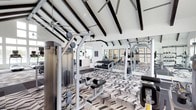 This fitness center is a thoughtfully designed workout facility that combines striking architectural details with a comprehensive selection of exercise equipment. The space is defined by its soaring vaulted ceiling adorned with bold, dark exposed beams that contrast beautifully against the crisp white walls, creating a dramatic and energizing atmosphere.

The flooring features a bold geometric chevron-patterned carpet that transitions to rubberized flooring in dedicated strength training zones, clearly delineating workout areas. Full-length mirrors line the walls, amplifying the sense of space and allowing users to monitor their form during exercise.

The strength training area is well-appointed with a multi-station cable machine, a dumbbell rack with a wide range of free weights, and several resistance and weight machines suited for various muscle groups. Cardio equipment includes multiple treadmills equipped with individual screens, elliptical trainers, and additional cardio machines, all positioned to take advantage of the floor-to-ceiling windows.

Natural light pours in through expansive windows, offering views of an outdoor pool area and the surrounding community, providing an inspiring backdrop for workouts. Wall-mounted flat-screen TVs are positioned throughout for entertainment. A separate glass-walled room is also visible, offering additional dedicated fitness or studio space. Ceiling fans ensure comfortable airflow throughout.