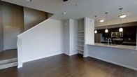This two-floor space showcases a thoughtfully designed open-concept layout that seamlessly connects the main living area to the kitchen. The living space features rich, dark hardwood flooring that extends throughout, complementing the clean, neutral-toned walls painted in a crisp light gray. A staircase leading to the upper floor is prominently positioned within the living area, adding architectural dimension to the space.

A built-in shelving unit is integrated into the wall, offering practical and decorative storage options. The kitchen is partially visible from the living area, separated by a peninsula counter finished with a granite-style countertop. Pendant lights hang above the counter, providing focused task lighting, while dark cabinetry and stainless steel appliances — including a full-sized refrigerator — contribute to a modern aesthetic. A ceiling fan with pull-cord accents is installed overhead in the living area. The overall ambiance is clean and contemporary, with a neutral color palette and functional design elements that create a cohesive and practical living environment across both floors.