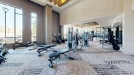 Located at 1881 N Rollins Road in Burlingame, California, this contemporary fitness center offers a thoughtfully designed, single-floor layout encompassing a generous workout space. The facility is divided into distinct functional zones, providing a comprehensive environment for a wide range of training needs.

The cardio area is lined with rows of treadmills, ellipticals, and stair climbers, each equipped with individual screens. Floor-to-ceiling windows flood this zone with abundant natural light and frame views of the surrounding outdoor greenery, creating an energizing atmosphere.

The strength training area features a variety of cable machines, selectorized equipment, and multi-functional training stations. A large flat-screen display is mounted on a teal accent wall, adding a dynamic focal point to the space. A dedicated free weights section offers a full rack of dumbbells alongside adjustable benches and floor mirrors for form monitoring.

A standout architectural feature is the soaring double-height ceiling in one section of the facility, accented by warm wood beam detailing and recessed cove lighting that lends a refined, upscale ambiance. The flooring throughout consists of a textured, patterned carpet tile in neutral tones. Additional amenities include a water fountain station near the entrance and a punching bag for boxing-style training. The overall design is modern and polished, reflecting a premium fitness facility.