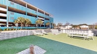Located at 1500 S Ocean Blvd in Myrtle Beach, South Carolina, this outdoor courtyard space captures the relaxed coastal character of the Grand Strand. The ground-level area is finished with lush artificial turf and features a variety of seating options, including lounge chairs, white picnic-style tables, and shaded nooks beneath a covered pergola structure — ideal for outdoor relaxation and social gatherings. Decorative breeze-block walls define the perimeter of the space, providing both privacy and a nod to mid-century coastal architectural style. Mature palm trees are planted throughout, adding natural texture and a distinctly tropical ambiance under the abundant South Carolina sunshine. The courtyard is nestled within the footprint of a multi-story building whose crisp white exterior is accented by striking teal-colored doors, lending the property a bold, retro-modern aesthetic. Upper-level balconies overlook the courtyard, creating a sense of enclosure and community. Stepping-stone pathways punctuate the turf, adding functional and visual detail to the layout. The space is open to the sky, bathed in natural light throughout the day, and benefits from the warm coastal climate characteristic of the Myrtle Beach area.
