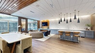 Located at 1 Franklin Town Blvd in Philadelphia's Logan Square neighborhood, this thoughtfully designed community lounge offers a refined yet welcoming atmosphere across a single, well-appointed floor. The space is divided into several functional zones that flow seamlessly into one another, anchored by warm hardwood flooring and a sophisticated palette of neutral tones, wood accents, and marble-inspired wall surfaces.

The lounge area is a standout feature, furnished with a plush curved sofa, leather armchairs, and a navy accent chair arranged around a central coffee table. A wall-mounted flat-screen TV is framed by warm wood paneling and a dramatic marble-effect accent wall, while a sleek console in muted blue-gray provides additional surface space. Large floor-to-ceiling windows flood the seating area with natural light and offer views of the surrounding greenery.

The kitchen and dining zone features a central island with bar seating beneath a striking cluster of black conical pendant lights with brass fixtures. The cabinetry continues the blue-gray palette against a full marble backsplash. Adjacent banquette and café-style seating provide casual dining options.

A separate corridor showcases a wood-slat ceiling, a gallery wall of framed artwork, and glass partition doors leading to additional community spaces. The bathroom is clean and accessible, with neutral tile flooring, grab bars, and modern fixtures. A full-length mirror and curated art touches complete the polished, community-centered design.