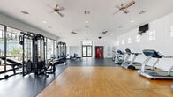 Located at 10881 Richmond Ave in Houston, TX, this well-equipped fitness center offers a generous, open exercise space designed to support a full range of workout routines. The single-floor facility features a thoughtful dual-flooring layout, combining warm hardwood in the open stretching and movement area with durable rubber matting beneath the equipment zones. Floor-to-ceiling windows line one wall, flooding the interior with abundant natural light and providing views of the surrounding outdoor area, creating an open and energizing atmosphere. The bright white walls and recessed lighting further enhance the sense of spaciousness throughout. Multiple ceiling fans are distributed across the ceiling, ensuring consistent airflow and comfort during workouts. The exercise room is outfitted with a variety of cardio machines, including treadmills, ellipticals, and a stationary bike, as well as multi-station strength training equipment, free weights, and benches to accommodate diverse fitness needs. A dedicated entry vestibule features double glass doors with transom windows, offering a welcoming transition into the space. A vending machine is conveniently tucked into an alcove near the entrance. The facility is monitored by a security camera system, providing a safe and well-maintained environment for users.