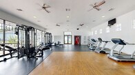 Located at 10881 Richmond Ave in Houston, TX, this well-equipped fitness center offers a generous, open exercise space designed to support a full range of workout routines. The single-floor facility features a thoughtful dual-flooring layout, combining warm hardwood in the open stretching and movement area with durable rubber matting beneath the equipment zones. Floor-to-ceiling windows line one wall, flooding the interior with abundant natural light and providing views of the surrounding outdoor area, creating an open and energizing atmosphere. The bright white walls and recessed lighting further enhance the sense of spaciousness throughout. Multiple ceiling fans are distributed across the ceiling, ensuring consistent airflow and comfort during workouts. The exercise room is outfitted with a variety of cardio machines, including treadmills, ellipticals, and a stationary bike, as well as multi-station strength training equipment, free weights, and benches to accommodate diverse fitness needs. A dedicated entry vestibule features double glass doors with transom windows, offering a welcoming transition into the space. A vending machine is conveniently tucked into an alcove near the entrance. The facility is monitored by a security camera system, providing a safe and well-maintained environment for users.