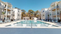 Located at 4830 Wescott Blvd in Summerville, South Carolina, this residential community combines modern architecture with inviting outdoor amenities. The exterior showcases contemporary multi-story buildings finished in classic grey and white siding, designed with a clean, cohesive aesthetic that complements the natural surroundings.

At the heart of the community lies a generously sized resort-style swimming pool, serving as the centerpiece of outdoor living. The pool area is thoughtfully designed with wide concrete decking and an abundance of lounge chairs, providing ample space for relaxation and recreation. Entry steps and handrails are conveniently positioned throughout the pool for easy access.

A charming covered gazebo structure anchors the far end of the pool area, offering a shaded gathering space ideal for socializing or unwinding outdoors. Decorative umbrellas add additional shading options along the poolside. The entire amenity area is framed by a backdrop of mature, tall trees that provide natural privacy and a tranquil, wooded atmosphere.

The community benefits from abundant natural light, with the open layout of the pool courtyard allowing sunlight to fill the space throughout the day. The well-maintained grounds and thoughtfully planned layout reflect a community designed with resident comfort and lifestyle in mind, making it an attractive destination in the greater Summerville area.