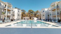 Located at 4830 Wescott Blvd in Summerville, South Carolina, this residential community combines modern architecture with inviting outdoor amenities. The exterior showcases contemporary multi-story buildings finished in classic grey and white siding, designed with a clean, cohesive aesthetic that complements the natural surroundings.

At the heart of the community lies a generously sized resort-style swimming pool, serving as the centerpiece of outdoor living. The pool area is thoughtfully designed with wide concrete decking and an abundance of lounge chairs, providing ample space for relaxation and recreation. Entry steps and handrails are conveniently positioned throughout the pool for easy access.

A charming covered gazebo structure anchors the far end of the pool area, offering a shaded gathering space ideal for socializing or unwinding outdoors. Decorative umbrellas add additional shading options along the poolside. The entire amenity area is framed by a backdrop of mature, tall trees that provide natural privacy and a tranquil, wooded atmosphere.

The community benefits from abundant natural light, with the open layout of the pool courtyard allowing sunlight to fill the space throughout the day. The well-maintained grounds and thoughtfully planned layout reflect a community designed with resident comfort and lifestyle in mind, making it an attractive destination in the greater Summerville area.