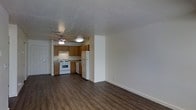 This single-floor residence offers a clean and functional layout designed for comfortable everyday living. The open-concept floor plan seamlessly connects the living area to the kitchen, creating a cohesive and practical space. Rich, dark wood-look flooring spans the living area, providing a warm and durable foundation that contrasts nicely with the bright white walls and textured ceiling overhead.

The kitchen is efficiently organized with warm honey-toned wooden cabinetry offering ample storage space. White appliances, including a gas range and a full-sized refrigerator, are neatly integrated into the layout, complemented by under-cabinet range ventilation. A ceiling fan with a built-in light fixture is centrally positioned, enhancing air circulation throughout the shared living and kitchen space.

The living area is generously proportioned, offering flexibility for a variety of furniture arrangements. Neutral wall tones throughout contribute to a bright and airy ambiance, making the space feel open and inviting. The textured ceiling adds a subtle visual detail that carries through the entire unit. Overall, the space presents a practical and well-maintained environment well-suited for comfortable, single-level living.