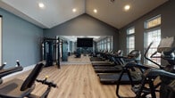 This modern fitness center is designed to support a comprehensive workout experience within a single open-plan floor. The space is defined by its striking vaulted ceilings, which lend an airy, expansive feel throughout the main exercise area. Walls are finished in a cool slate-gray tone, complemented by warm wood-look flooring that runs throughout, creating a balanced and motivating atmosphere.

The main workout floor is outfitted with a row of treadmills and stationary bikes for cardio training, alongside a multi-function cable and strength machine system for resistance work. A full rack of dumbbells lines one wall beneath a large flat-screen television, making it easy to follow along with workout content. A floor-to-ceiling wall mirror spans a significant portion of the room, visually expanding the space and allowing users to monitor their form.

Natural light flows in through multiple windows, including transom windows positioned near the roofline, further enhancing the open and energetic environment. Recessed lighting provides consistent illumination throughout.

A separate studio room, accessible via glass sliding doors, offers a more focused training or stretching area. This secondary space includes a small workstation, a compact beverage refrigerator, and mirrored walls, making it suitable for personal training or group fitness sessions. Together, these areas form a thoughtfully designed, fully functional fitness facility.