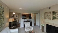 This single-floor residence offers a thoughtfully designed open-concept layout that seamlessly connects the main living areas. The living room serves as the heart of the home, featuring plush carpeting, neutral-toned walls, and ample space for comfortable furnishings. A ceiling fan overhead provides both function and a finishing touch to the room's understated aesthetic. Adjacent to the living area, the dining space accommodates a full dining set and is accented by decorative wall art, creating a warm and inviting atmosphere for gatherings. The kitchen is positioned at the rear of the open floor plan and features dark wood cabinetry, black appliances including a microwave and refrigerator, and a peninsula counter that doubles as a bar or serving area. The peninsula provides a natural transition point between the kitchen and living spaces, enhancing the open flow of the home. A hallway visible from the main living area suggests additional rooms beyond, including what appears to be access to a bathroom or bedroom. The overall color palette of warm beiges and browns, combined with the open layout and consistent flooring transitions, gives the space a cohesive and welcoming character throughout.