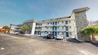 This multi-story building presents a classic Florida-style exterior characterized by a clean white facade accented with teal doors and railings that run along each level's covered walkway. The structure spans three floors, with each level featuring an open-air corridor that provides direct access to individual units. Private balconies lined with white railings extend from the upper floors, offering outdoor space for each unit. A notable architectural detail is the prominent stone accent tower on one end of the building, adding texture and visual contrast to the otherwise smooth exterior walls. The roofline features a traditional pitched design with shingle cladding that complements the overall coastal aesthetic. Ground-level units benefit from direct parking lot access, while upper-floor units are reached via exterior staircases. The paved parking area directly fronts the building, providing convenient off-street parking. Palm trees flanking the property reinforce the warm-weather, coastal character of the building. The exterior is well-maintained, with security cameras visible along the facade. Interior images were not sufficiently lit to provide detailed information about the interior spaces.