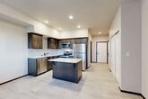 Located in Arlington, Washington, this thoughtfully designed single-floor unit offers a compact yet functional layout that maximizes every square foot. The heart of the space is an open-concept area combining the kitchen, dining room, and living room, creating a seamless flow ideal for everyday living. The kitchen stands out with rich dark-stained cabinetry, a classic white subway tile backsplash, light quartz countertops, and a full suite of stainless steel appliances including a refrigerator, electric range, microwave, and dishwasher. A central island provides additional prep space and casual seating. Light wood-look flooring runs throughout the unit, complementing the neutral, warm color palette of the walls and trim. A dedicated den offers flexibility as a home office, reading nook, or additional storage space. The combined bathroom and laundry room is efficiently designed, featuring a dark-stained vanity with a quartz countertop, a stacked washer and dryer, and clean modern finishes. Two closets provide ample storage, and a welcoming entrance sets the tone for the unit's clean, contemporary aesthetic. A window in the living area frames views of surrounding greenery, bringing natural light and a connection to the outdoors into the space.