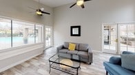 This well-appointed single-floor space combines modern comfort with clean, contemporary design throughout. The heart of the living area is a striking fireplace framed by a crisp white mantel and beige tile surround, anchoring the room with warmth and visual interest. Light-toned wood-look flooring runs throughout, complementing the neutral warm-gray walls and bright white trim and baseboards. A gently curved bay window area floods the living space with natural light, offering views of the surrounding outdoor greenery and pool area. Soaring ceilings with ceiling fans add to the open, airy feel of the main living area, which comfortably accommodates multiple seating arrangements. Large windows with horizontal blinds allow for ample light control while maintaining a connection to the outdoors. A set of glass doors provides direct access to the exterior. The kitchen is thoughtfully designed with granite countertops, white shaker-style cabinetry, a double-basin sink beneath a bright window, and built-in appliances, offering both practicality and style. The overall ambiance of the space is bright, clean, and inviting, with a cohesive neutral palette that creates a calm and versatile environment throughout.