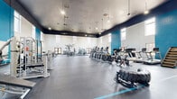 Located at 2707 S Rutherford Blvd in Murfreesboro, TN, this single-floor fitness facility offers an impressive and well-equipped space designed to support a wide range of workout disciplines. The centerpiece is a vast main exercise floor characterized by soaring ceilings, bold teal accent walls, and rows of clerestory windows that flood the space with natural light. The dark ceiling trim contrasts sharply with the bright white walls, lending the gym a modern, energetic aesthetic. The main floor is outfitted with an extensive array of equipment, including Matrix cardio machines such as treadmills, ellipticals, and stair climbers, alongside cable systems, weight racks, benches, squat racks, and functional training implements including a heavy tire. Full-length mirrors line portions of the walls, enhancing both the sense of space and functionality for strength training. A dedicated studio room features luxury vinyl flooring, wall-mounted exercise mats, and a large mirror wall, making it ideal for group fitness, yoga, or stretching. An additional open room with light flooring and neutral walls provides flexible multipurpose space. The hallway area is appointed with a large multi-cubby wooden shelving unit for personal storage, alongside access to restroom facilities. Consistent recessed and fluorescent lighting throughout ensures a bright, functional environment across all areas.