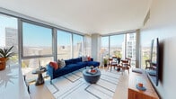 Located at 100 S Grand Avenue in the heart of Downtown Los Angeles, this single-floor residence offers a refined urban living experience defined by expansive floor-to-ceiling windows that frame panoramic city skyline views, including sightlines toward the Walt Disney Concert Hall and surrounding high-rises. Natural light floods the open-plan living and dining area, where light hardwood flooring and clean-lined architecture create a contemporary aesthetic throughout.

The living space is generously proportioned, accommodating both a lounge and dining area side by side, with sheer drapery softening the abundant sunlight. The kitchen features sleek, flat-front cabinetry in a warm white finish, a striking stone backsplash, quartz countertops, stainless steel appliances, and a large island, all positioned adjacent to city views.

The bedroom offers a calm retreat with textured accent wallcovering and hardwood floors. Two bathrooms serve the residence: one with warm wood cabinetry, backlit mirrors, and stone-tiled flooring, and another featuring a soaking tub with glass enclosure and large-format tile. A welcoming entry hallway with a round mirror and a flexible secondary sitting room complete this well-appointed urban residence.