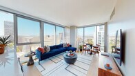 Located at 100 S Grand Avenue in the heart of Downtown Los Angeles, this single-floor residence offers a refined urban living experience defined by expansive floor-to-ceiling windows that frame panoramic city skyline views, including sightlines toward the Walt Disney Concert Hall and surrounding high-rises. Natural light floods the open-plan living and dining area, where light hardwood flooring and clean-lined architecture create a contemporary aesthetic throughout.

The living space is generously proportioned, accommodating both a lounge and dining area side by side, with sheer drapery softening the abundant sunlight. The kitchen features sleek, flat-front cabinetry in a warm white finish, a striking stone backsplash, quartz countertops, stainless steel appliances, and a large island, all positioned adjacent to city views.

The bedroom offers a calm retreat with textured accent wallcovering and hardwood floors. Two bathrooms serve the residence: one with warm wood cabinetry, backlit mirrors, and stone-tiled flooring, and another featuring a soaking tub with glass enclosure and large-format tile. A welcoming entry hallway with a round mirror and a flexible secondary sitting room complete this well-appointed urban residence.
