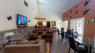 Located at 1651 S Dobson Rd in Mesa, Arizona, this single-floor community clubhouse offers a well-appointed gathering space designed for comfort and functionality. The main lounge area commands attention with soaring vaulted ceilings and an abundance of natural light streaming in through clerestory windows positioned high on the walls. A striking stone accent wall serves as the focal point of the lounge, anchoring a wall-mounted flat-screen television flanked by decorative sconce lighting. Generous seating arrangements with plush sofas and armchairs are arranged over a colorful area rug, creating a warm and inviting atmosphere. Adjacent to the lounge, a dining area provides ample table and chair seating, ideal for community gatherings. The fully equipped kitchen features rich wood cabinetry, dark granite countertops, black appliances including a range, microwave, and French door refrigerator, along with a double-basin sink beneath a natural-light window. A dedicated community workstation nook with a desktop computer and bulletin board offers a practical resource for residents. French doors with glass panes connect the interior spaces, enhancing the open and airy feel throughout. Dark wood-look flooring ties the entire space together, lending a cohesive and polished aesthetic throughout the clubhouse.