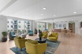 The Lounge at 10568 Gateway Promenade is a thoughtfully designed community business center located within a residential complex in El Monte, California. Spanning a generous single floor, this contemporary space is defined by an open, airy layout bathed in natural light from expansive floor-to-ceiling sliding glass doors that frame direct views of the outdoor pool and courtyard beyond.

The main lounge area showcases a modern aesthetic with light hardwood flooring, clean white ceilings with recessed lighting, and a neutral color palette accented by vibrant furnishings. Bold mustard-yellow lounge chairs and teal accent seating are arranged atop teal-toned area rugs, creating inviting conversation clusters throughout the space. Lush potted plants add a refreshing natural element to the interior.

A dedicated dining area features a communal high-top table with modern wooden stools and vivid framed artwork on the walls, alongside a warm wood-grain credenza that provides additional functional storage. A separate, more intimate dining nook offers a marble-topped bistro table with stylish bar stools beneath a sleek pendant light — ideal for focused work or casual meetings.

Colorful curated artwork adorns the walls throughout, contributing to an energetic yet welcoming atmosphere. The seamless connection between the lounge, dining zones, and outdoor views makes this a versatile and inspiring shared space.