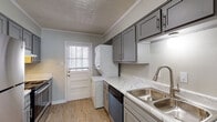 This single-floor, two-bedroom, one-and-a-half-bath home in Memphis, TN offers a thoughtfully updated interior with a clean, modern aesthetic. The kitchen serves as a standout feature, showcasing gray shaker-style cabinetry with brushed hardware, marble-look quartz countertops, a stainless steel double-basin sink, and a full suite of appliances including a dishwasher, stainless refrigerator, and range. A stacked washer and dryer unit is conveniently integrated into the kitchen area, and a rear entry door with a window brings in additional natural light. Crown molding adds a polished finish throughout the home. Light wood-look vinyl plank flooring flows consistently across the living areas and both bedrooms, complementing the soft gray wall tones. The living area connects openly to the kitchen via a hallway, creating a functional layout. Both bedrooms are generously sized, each featuring windows with blinds that allow ample natural light. The full bathroom is finished with classic white subway tile in the tub/shower surround, marble-look floor tile, and brushed bronze fixtures. The half bath offers a white vanity with a clean countertop and a framed mirror, providing a crisp, updated look.