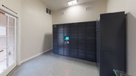 This single-floor mail and package room at Arbor Pointe is a dedicated, functional space designed to streamline parcel and mail retrieval for residents. The room is accessed through a glass-paneled exterior door equipped with a secure lock, offering a clear view of the surrounding outdoor walkway and courtyard area. A "Please Keep Door Closed" sign underscores the secured nature of the entry point.

Inside, the space features speckled epoxy flooring throughout, lending a clean and durable finish that is easy to maintain. The walls are finished in a neutral warm beige tone with white baseboards, creating a bright and orderly environment. Natural light filters in through a window fitted with white horizontal blinds, supplementing the overhead fluorescent lighting.

A prominent feature of the room is the large Amazon Hub smart locker system, which spans a significant portion of one wall. This multi-compartment, self-service parcel locker allows residents to securely receive and retrieve packages at their convenience. Adjacent to the lockers, a bank of traditional individual mailboxes labeled "Arbor Pointe – All Units" provides organized mail storage for all residents.

A decorative glass-top console table with an ornate gold metal base adds a touch of elegance to the otherwise utilitarian space. A security camera is also visible, ensuring the area remains monitored for resident safety.