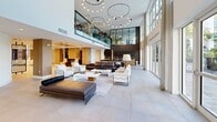 Located at 10900 Legacy Gateway Cir in Fort Myers, Florida, this modern community clubhouse delivers an impressive sense of arrival and openness across its single-floor layout. The grand entrance foyer welcomes visitors with soaring double-height ceilings, large-format porcelain tile flooring, and striking red vertical rod sculptures that serve as bold architectural focal points. Floor-to-ceiling glass walls and expansive windows flood the interior with natural light while framing lush views of swaying palm trees, a resort-style pool, and the surrounding landscape.

The main living area is thoughtfully appointed with a mix of rich leather and upholstered seating arrangements, anchored by dramatic multi-tiered circular chandeliers that cascade elegantly from the lofted ceiling. A glass-railed mezzanine overlooks the main floor, adding architectural depth and visual interest. The kitchen and lounge areas feature warm wood cabinetry and contemporary finishes, while a dedicated living room offers a more intimate setting with a textured dark accent wall, curated artwork, and plush furnishings. Additional zones include a media area with a wall-mounted display set against natural wood plank paneling. Throughout, the design balances bold artistic elements with a refined, welcoming ambiance.