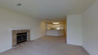 This single-floor space is designed with an open-concept layout that seamlessly connects the living area and kitchen. The living area is anchored by a classic brick fireplace fitted with a glass-front gas insert, adding both warmth and a focal point to the room. Neutral-toned carpeting extends throughout the space, contributing to a calm and cohesive aesthetic.

The kitchen is positioned toward the rear of the open floor plan and features white cabinetry that extends along the walls, providing ample storage. A curved peninsula counter serves as a natural divider between the kitchen and living area, offering additional workspace and a casual dining option. Recessed lighting and a fluorescent fixture illuminate the kitchen effectively, while a track lighting unit in the transition zone adds supplemental overhead light. White appliances, including what appears to be a refrigerator and additional built-in units, are integrated into the cabinetry layout.

An additional doorway visible beyond the kitchen suggests access to further areas of the unit. The overall palette of white walls, light cabinetry, and neutral carpet creates a clean, understated interior environment.