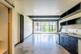 This single-floor space showcases a striking industrial-modern aesthetic defined by exposed black steel beams, corrugated metal ceilings, and polished concrete floors throughout. The open main area is bathed in natural light streaming through large floor-to-ceiling windows, offering views of surrounding greenery and creating an airy, expansive atmosphere. Bold diagonal steel bracing adds a dramatic architectural element, further emphasizing the industrial character of the space.

The kitchen is thoughtfully designed with sleek dark cabinetry, dark countertops, stainless steel appliances including a full-size refrigerator, electric range, and microwave, along with a stainless steel sink. A sliding barn-style door opens to a built-in closet with shelving, providing practical storage near the main entrance.

The bathroom features a glass-enclosed walk-in shower with tile surround and a window for natural light, complemented by a stacked washer and dryer unit — a convenient in-unit laundry solution. Recessed lighting throughout provides consistent illumination, while an industrial-style pendant fixture adds a decorative touch to the main living area. The combination of raw materials, open layout, and abundant natural light gives this space a versatile, contemporary character well-suited to a variety of uses.