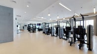 This contemporary fitness center offers a spacious, single-floor layout thoughtfully designed to accommodate a comprehensive range of exercise equipment. The open floor plan allows for generous spacing between machines, ensuring a comfortable and unobstructed workout environment.

The facility is outfitted with an extensive selection of strength training machines, power racks, and functional training stations, providing options for users of all fitness levels. Rows of equipment are arranged in an organized, easy-to-navigate configuration throughout the space.

Overhead, a combination of recessed lighting and stylish pendant fixtures illuminates the facility with bright, even light. The high, coffered white ceiling amplifies the sense of openness and airiness throughout. Large windows along the perimeter flood the space with natural light and offer views of the surrounding greenery, creating an inviting atmosphere.

The flooring features a durable, light-toned surface well-suited for high-traffic fitness use, while the neutral gray and white color palette contributes to a clean, modern aesthetic. Mounted flat-screen televisions are positioned throughout the space to keep users engaged during their workouts. The overall ambiance is professional, energizing, and welcoming — reflecting a fitness center built for both performance and comfort.