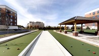 This outdoor recreational space offers a well-appointed bocce ball facility designed for both casual play and organized competition. The courts are finished with a smooth, green synthetic surface framed by clean concrete borders, providing a professional and consistent playing experience. Multiple lanes run parallel, accommodating several groups of players simultaneously.

Adjacent to the courts stands a handsome covered pavilion, constructed with warm wood timber columns resting on classic brick bases and topped with a dark, pitched roof. The structure provides a shaded gathering area, ideal for spectators or players taking a break between matches.

The surrounding landscape is thoughtfully maintained, with neatly trimmed green lawns and a variety of trees that offer a natural, park-like setting. The open layout allows for generous natural light and unobstructed views of the sky, creating a bright and welcoming atmosphere. A multi-story building is visible in the background, suggesting this amenity is part of a larger hospitality or residential complex. The overall design balances function and aesthetics, making this space an inviting destination for outdoor recreation and social gathering.
