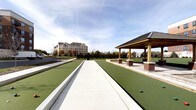 This outdoor recreational space offers a well-appointed bocce ball facility designed for both casual play and organized competition. The courts are finished with a smooth, green synthetic surface framed by clean concrete borders, providing a professional and consistent playing experience. Multiple lanes run parallel, accommodating several groups of players simultaneously.

Adjacent to the courts stands a handsome covered pavilion, constructed with warm wood timber columns resting on classic brick bases and topped with a dark, pitched roof. The structure provides a shaded gathering area, ideal for spectators or players taking a break between matches.

The surrounding landscape is thoughtfully maintained, with neatly trimmed green lawns and a variety of trees that offer a natural, park-like setting. The open layout allows for generous natural light and unobstructed views of the sky, creating a bright and welcoming atmosphere. A multi-story building is visible in the background, suggesting this amenity is part of a larger hospitality or residential complex. The overall design balances function and aesthetics, making this space an inviting destination for outdoor recreation and social gathering.
