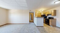This single-floor, two-bedroom unit offers a practical and well-organized layout totaling approximately 710 square feet. The open-concept living area flows seamlessly into the kitchen, which is defined by a peninsula with a dark granite-style countertop — ideal for casual dining or additional prep space. The kitchen is equipped with oak-toned cabinetry, black appliances including a refrigerator, range, and dishwasher, and durable tile flooring with track lighting overhead. The living area features neutral carpet and a textured ceiling, creating a clean, understated aesthetic throughout.

The primary bedroom is a generously sized, carpeted room with natural light from a large double-hung window. It is complemented by a spacious walk-in closet outfitted with wire shelving on multiple walls, offering ample storage capacity. The second bedroom is also carpeted and features its own window with views of the surrounding neighborhood. Both bedrooms maintain a simple, neutral color palette with warm wood-toned baseboards and doors.

Two hallways connect the private and shared spaces efficiently, adding to the functional flow of the unit. Overall, the space presents a clean, move-in ready design with consistent finishes throughout.