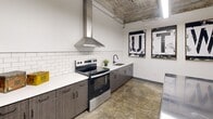 This single-room kitchen space showcases a striking blend of industrial and modern design elements. The exposed concrete ceiling, complete with visible structural elements and overhead fluorescent lighting, sets a raw, urban tone throughout the space. Crisp white walls contrast with the textured ceiling, creating a clean and functional environment.

The kitchen is equipped with a full suite of stainless steel appliances, including a French door refrigerator, an electric range, and a prominent stainless steel wall-mounted range hood that serves as a bold focal point. The cabinetry features a dark wood-grain finish with matte black hardware, paired with white countertops and a classic white subway tile backsplash that adds a timeless touch. A stainless steel sink with a matte black faucet completes the cooking and prep area.

At the center of the room, a large stainless steel prep table with wooden stools provides additional workspace and a gathering point, reinforcing the kitchen's professional and utilitarian character. A wall-mounted mini-split HVAC unit ensures climate control, while polished concrete floors tie the industrial aesthetic together seamlessly.

Decorative black-and-white typographic art pieces adorn the walls, adding a curated, creative touch to the otherwise utilitarian space. A gray steel entry door leads out of the room, completing this well-appointed, efficiently designed kitchen.