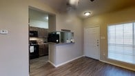 This single-story residence offers a practical and cohesive layout designed for comfortable everyday living. The entryway greets visitors with a classic paneled white door and opens directly into the main living area, which features warm hardwood-style flooring that flows seamlessly throughout the space. A ceiling fan overhead provides both function and a subtle design accent.

The kitchen is open to the main living area and is equipped with dark wood cabinetry that provides ample storage, complemented by granite-style countertops that add a touch of refinement. Black appliances, including a range and microwave, lend a uniform, modern aesthetic to the cooking space. A double sink sits neatly beneath the counter, and the layout allows for efficient meal preparation.

A half-wall pass-through counter connects the kitchen to the adjacent area, enhancing the open feel of the floor plan while offering a casual space for dining or entertaining. The textured ceiling and neutral warm-toned walls contribute to an inviting ambiance throughout. The overall design prioritizes functionality and flow, making the space well-suited for daily living while maintaining a clean and cohesive interior style.